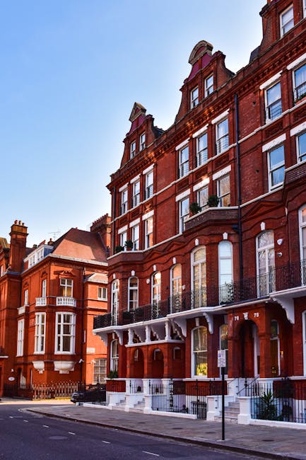 Photograph of the Royal Albert Hall in Kensington, London, showcasing its circular brick facade with decorative arches and large windows. The building is surrounded by a well-maintained grassy area wi