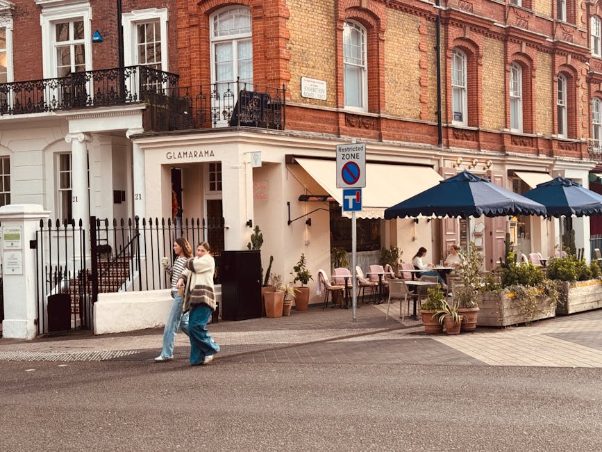 Exterior view of a building with a sign reading 'Glamarama' above the entrance, featuring a white facade with black iron railings and large glass windows. The sidewalk in front has outdoor seating arrangements with pink chairs and large navy blue umbrellas shading the tables. There are potted plants and flowers along the edge of the seating area, adding greenery. Two women are walking on the sidewalk, engaged in conversation. The building's upper floors showcase red brick walls with white-framed windows and decorative iron balconies, illuminated by soft daylight, suggesting a clean and inviting atmosphere. The scene is captured from street level, highlighting the clean, well-maintained storefront and outdoor dining area, consistent with the services offered by Cleaner Kensington for domestic and commercial cleaning.