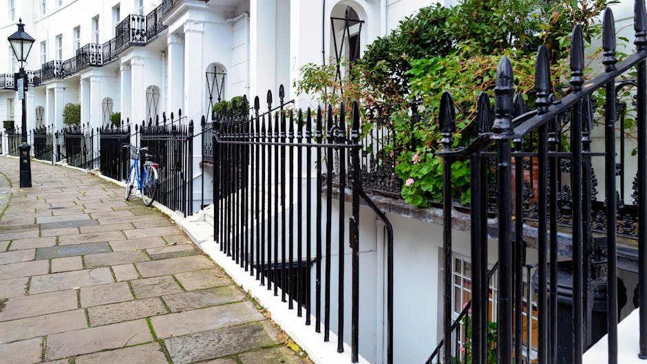 A paved sidewalk running alongside a row of white residential buildings with black wrought iron railings. The pavement consists of rectangular stone slabs, some showing signs of weathering, arranged in a neat pattern. A blue bicycle is parked against one of the railings, partially visible. Green leafy plants and pink flowering vines grow along the fence, adding a touch of greenery. The buildings have large windows with black metal frames and some feature decorative ironwork balconies, illuminated by natural light that highlights the cleanliness of the surfaces and the well-maintained appearance of the exterior. Cleaner Kensington's logo and branding are associated with the scene, emphasizing quality domestic and commercial cleaning services, although not directly visible in this image.
