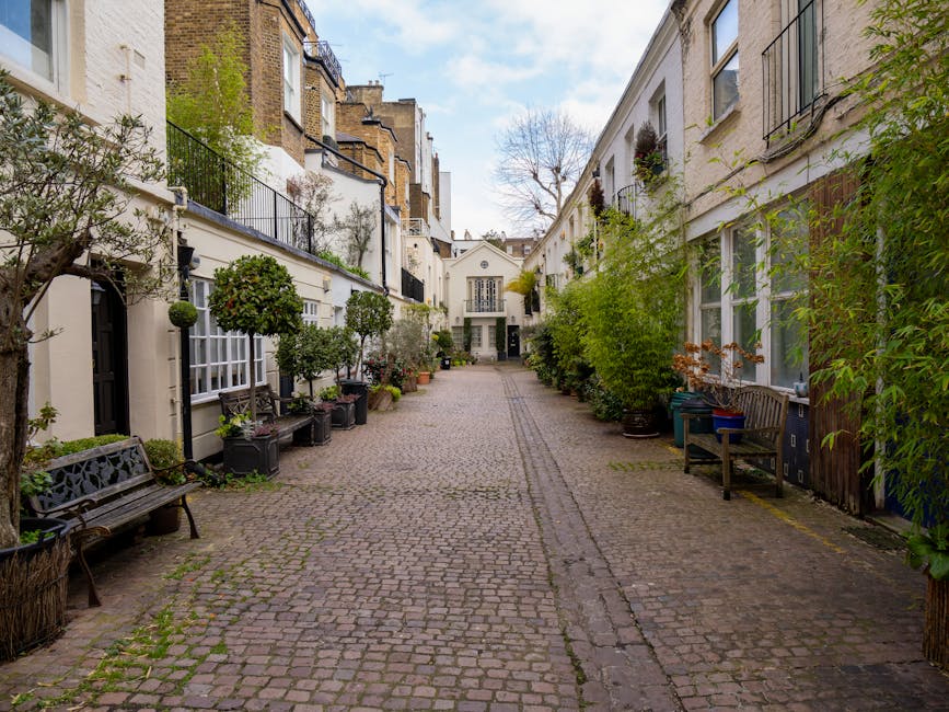 A narrow residential alley lined with white and beige buildings on both sides, featuring black wrought iron balconies and window frames. The cobblestone pathway is clean and dry, bordered by neatly arranged potted plants and trees in dark containers, some with green foliage and others with blossoms. Wooden benches are placed along the walls, and the lighting is natural with a partly cloudy sky overhead. The scene reflects well-maintained outdoor spaces typical of UK townhouses, with no visible debris or dirt, suggesting regular surface cleaning and garden maintenance by firms such as Cleaner Kensington.