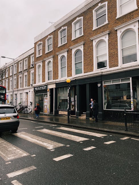 A quiet, cobblestone street in Kensington lined with red-brick residential and commercial buildings featuring large windows and small balconies. The street is lined with black bollards and vintage-sty