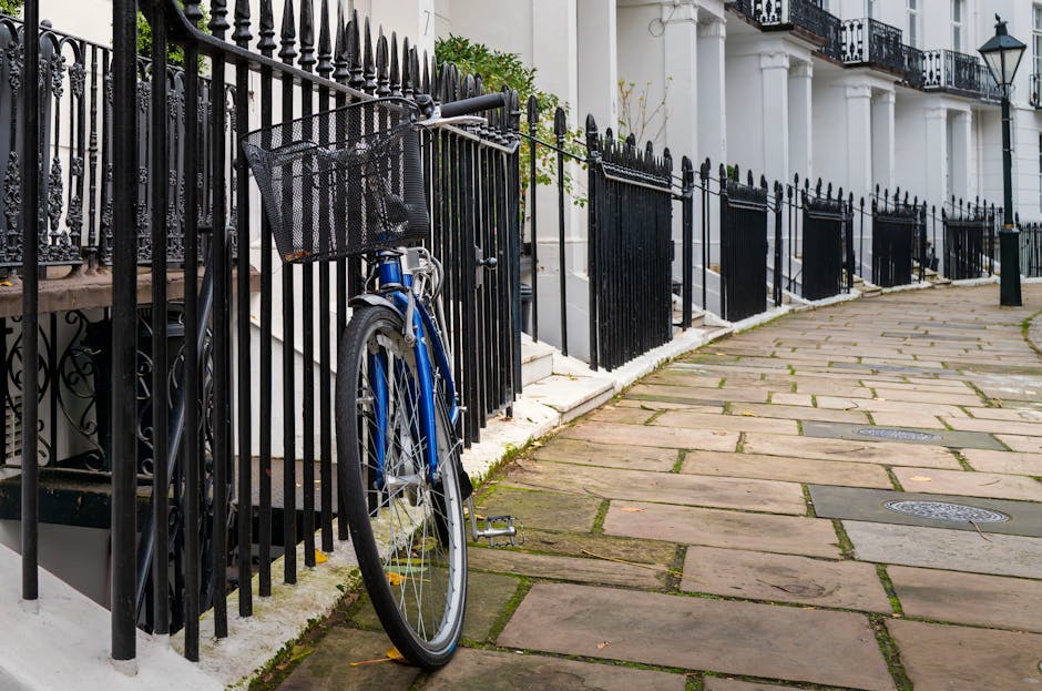A sidewalk in Kensington W8 with large rectangular stone paving slabs, some with visible moss or dirt in the joints, bordered by black wrought iron fencing with decorative finials. A blue bicycle with a black basket is leaning against the fence near the small staircase leading to a white building with classical columns and black wrought iron balcony railings. The scene is well-lit with natural daylight, highlighting the clean, well-maintained surfaces and tidy appearance of the area, exemplifying surface maintenance and exterior cleaning, with attention to detail in the cleanliness of the pavement and fencing, as provided by Cleaner Kensington.