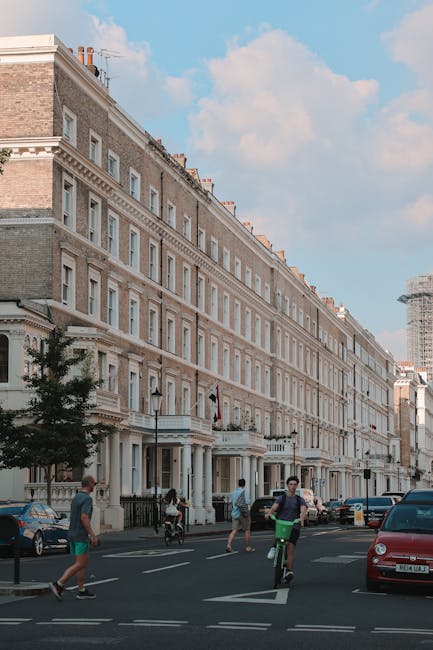 The image depicts a narrow residential street paved with cobblestones, flanked by white and beige terraced houses with small front gardens decorated with potted plants. A leafless tree stands prominently in the background against a cloudy sky. On the right side, a tall stone wall covered partly with green ivy separates the street from neighboring properties. The scene is well-lit with natural daylight, emphasizing the clean, orderly appearance of the street and the vibrant greenery, adding to the calm atmosphere typical of Kensington. Cleaner Kensington specialises in surface cleaning and deep cleaning services, ensuring the hygiene and pristine condition of such residential areas as shown in the image.