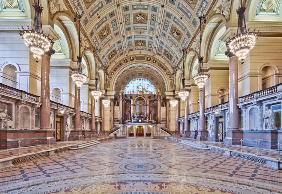 An interior view of the Royal Albert Hall showcasing an opulent hall with a high, ornate ceiling decorated with detailed moldings and painted panels. Grand, cylindrical marble columns with decorative capitals line the sides of the space, supporting large, elegant chandeliers hanging from the ceiling. The polished wooden and marble floors are spotless, reflecting the lighting and enhancing the room's grandeur. At the far end, a stained glass window filters natural light into the hall, complementing the artificial lighting from the chandeliers. Gold and cream-colored accents, along with intricate wall sculptures and notices, add to the luxurious atmosphere. Cleaner Kensington has maintained the space's pristine condition, evident in the gleaming surfaces and meticulously arranged area, suitable for formal events or performances.