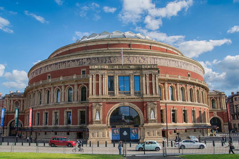 A large, circular historic building with a domed roof, constructed with red brick and stone accents, featuring a series of arched windows and statues along its facade. The front entrance displays a sign reading 'Royal Albert Hall' beneath a large, arched doorway. In front of the building, a street scene includes cars, bicycles, and pedestrians, with a clear blue sky overhead. The image reflects the exterior surface cleaning and maintenance typical of professional cleaning services like those offered by Cleaner Kensington, emphasizing the building's pristine appearance and structural details.