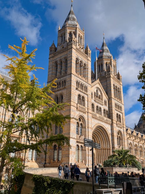A large, historic church with multiple tall towers featuring pointed spires, constructed from light-colored stone with detailed architectural carvings and arched windows. The building is set against a bright blue sky with some scattered clouds and is partially framed by green trees. In front of the church, there is a paved area with several people walking and gathering near the entrance. The overall scene highlights the intricate stonework and grand scale of the church, which is located in a busy urban setting. This image exemplifies the architecture that might require routine surface cleaning and maintenance, and is referenced in relation to same day emergency cleaning services offered by Cleaner Kensington for heritage and commercial properties.