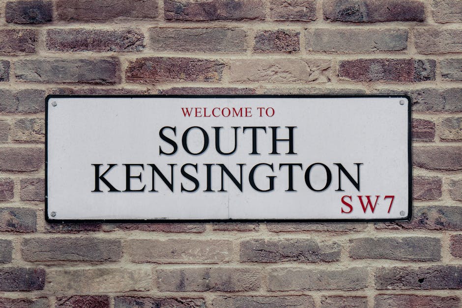 A rectangular metal street sign mounted on a red brick wall, displaying the text 'Welcome to South Kensington SW7' in black and red lettering on a white background, with visible rivets at each corner. The brick wall consists of uniformly sized bricks with mortar joints, providing a textured surface behind the sign. The sign's surface appears clean and well-maintained, indicating a focus on clear signage and exterior presentation, consistent with professional cleaning standards promoted by Cleaner Kensington for domestic and commercial environments.