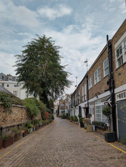 A quiet, cobblestone street in Kensington lined with red-brick buildings featuring large windows and small balconies, some with black wrought-iron railings. The street is shaded by a lamppost and gree