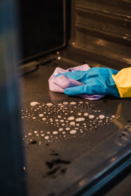 Close-up of a person's hand wearing a blue rubber glove and a yellow sleeve, using a purple cloth to clean the black, stainless steel surface inside an oven. The surface shows small soap bubbles and streaks, indicating an ongoing deep cleaning process. The oven interior features a textured silver wall with ridges, and the lighting highlights the reflective nature of the metal. This image illustrates surface cleaning and sanitisation activities undertaken by Cleaner Kensington as part of their domestic cleaning services for ovens in South Kensington SW7, emphasizing hygiene and maintenance of kitchen appliances.