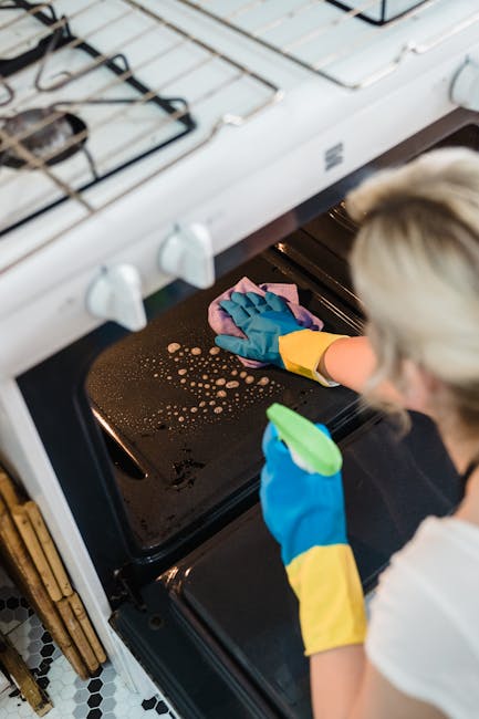 Close-up of a person's hand wearing a blue rubber glove and a yellow sleeve, using a purple cloth to clean the black, stainless steel surface inside an oven. The surface shows small soap bubbles and streaks, indicating an ongoing deep cleaning process. The oven interior features a textured silver wall with ridges, and the lighting highlights the reflective nature of the metal. This image illustrates surface cleaning and sanitisation activities undertaken by Cleaner Kensington as part of their domestic cleaning services for ovens in South Kensington SW7, emphasizing hygiene and maintenance of kitchen appliances.